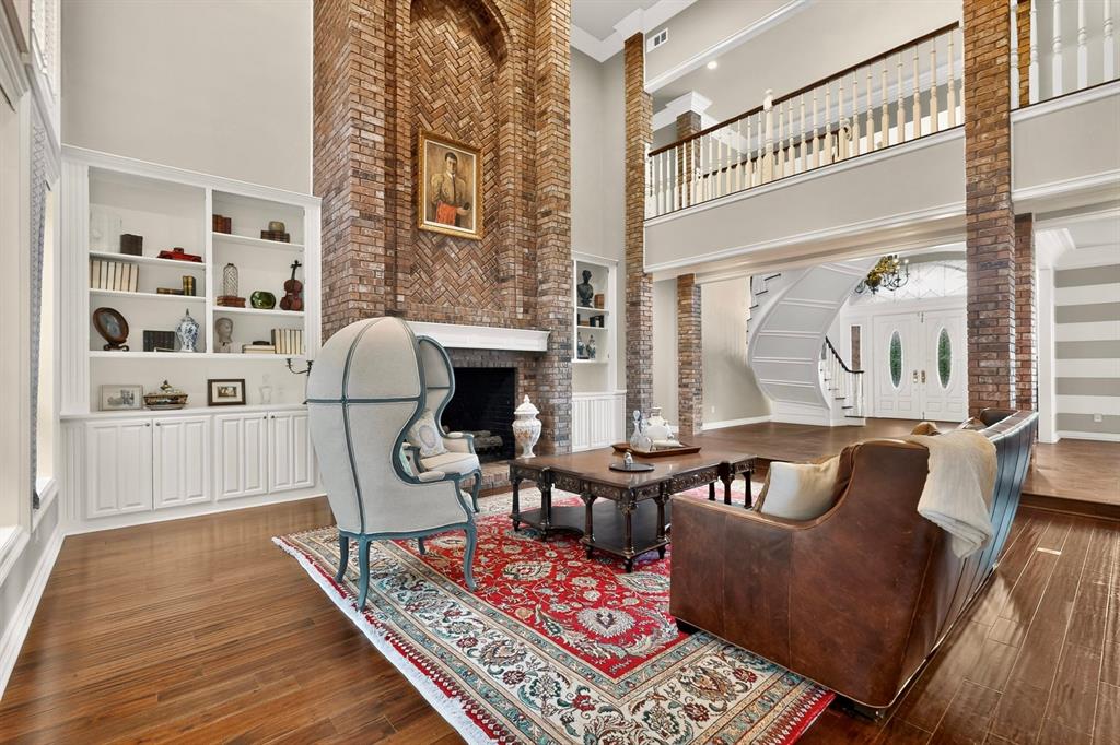 Two-story grand hall with floor-to-ceiling brick fireplace, herringbone-patterned masonry detail, built-in shelving, wood-finish flooring, and a curved staircase with a traditional railing