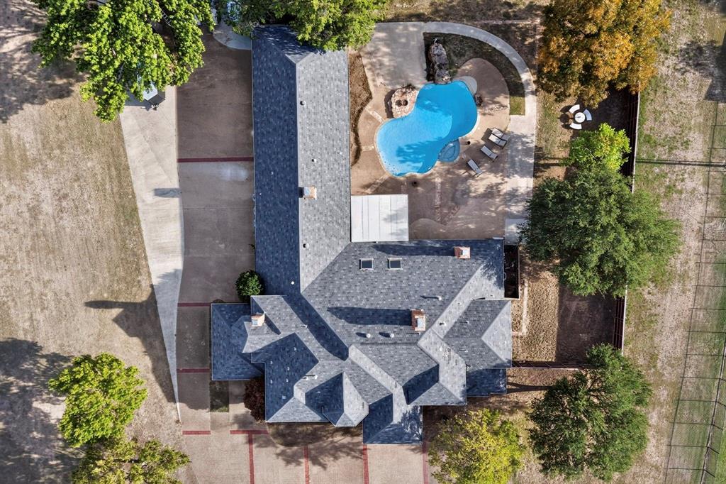 Aerial perspective of the property featuring a sprawling asphalt-shingle roof, a freeform swimming pool with integrated spa, an extensive concrete driveway, and mature landscaping on a large lot