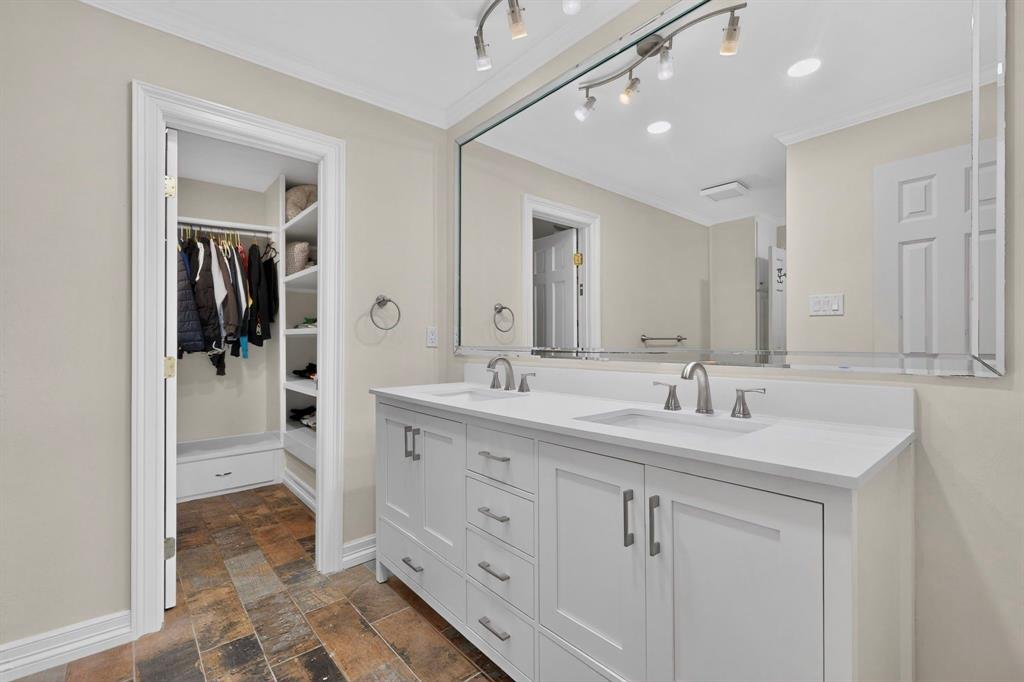 Double-sink vanity with white cabinetry, light-toned stone-finish countertop, brushed metal hardware, large wall-mounted mirror, and tile flooring with integrated shelving within the closet area