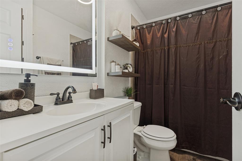 White vanity with a solid-surface countertop, under-mount sink, matte black hardware, integrated LED-backlit mirror, and tiered wood-finish floating shelving