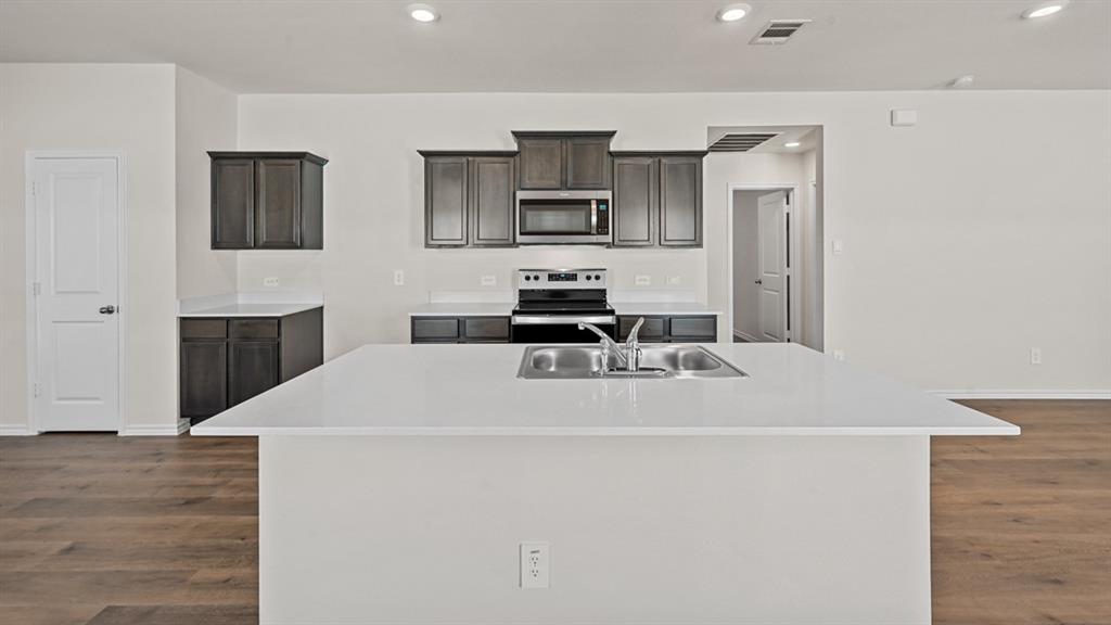 Spacious kitchen with a central island featuring an integrated stainless steel sink, dark wood-finish cabinetry, light-toned countertops, and wood-finish flooring