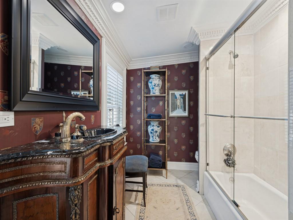 Custom vanity with ornate wood detailing, stone countertop, integrated sink, and wall-mounted mirror, featuring crown molding, tile flooring, and a shower-tub combination with a glass enclosure