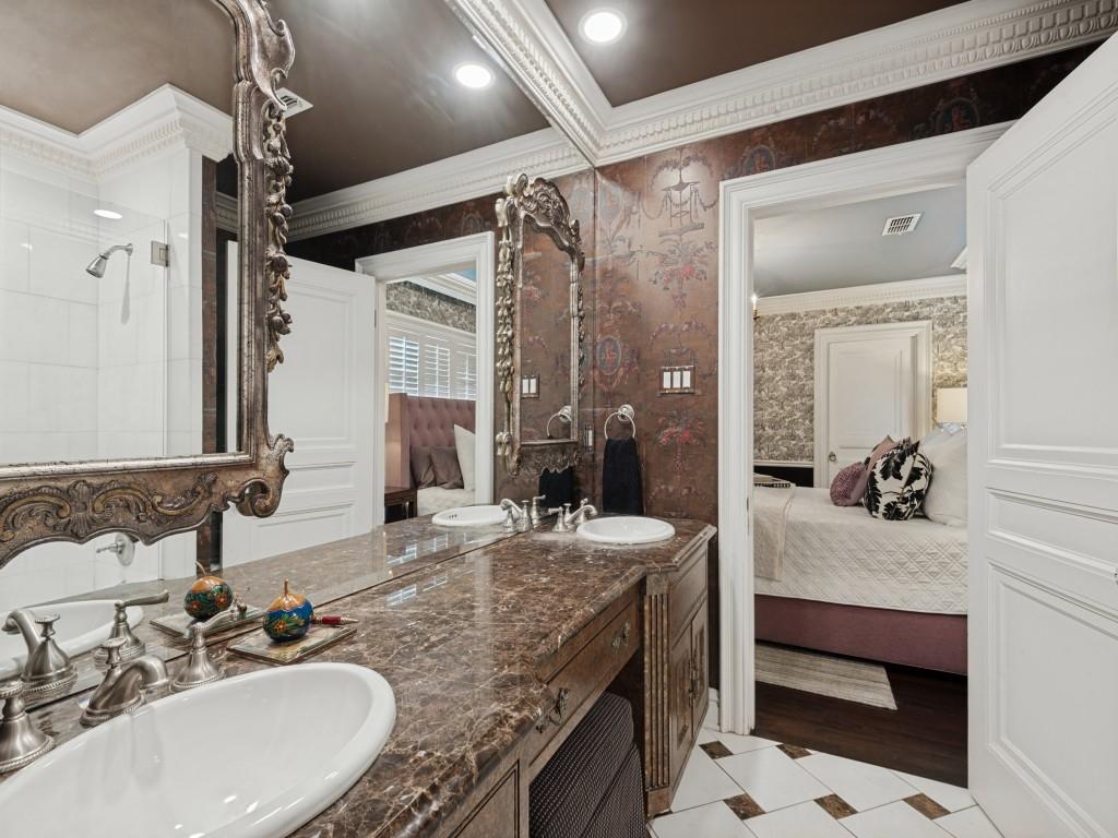 Double vanity featuring stone countertops with dual white drop-in sinks, ornate wall-mounted mirrors, custom cabinetry, and patterned wall coverings complemented by detailed crown molding and tile flooring