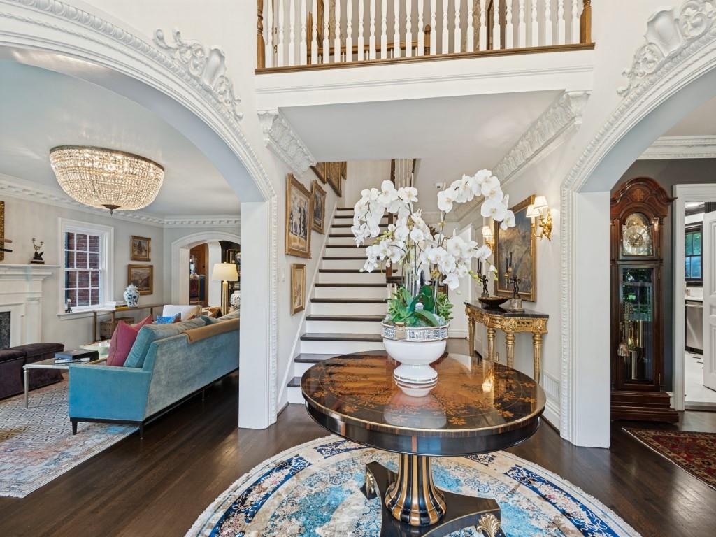 Grand foyer featuring intricate crown molding, arched entryways, dark wood-finish flooring, a crystal chandelier, and a staircase with a wooden handrail and balusters Grand foyer featuring intricate crown molding, arched entryways, dark wood-finish flooring, a crystal chandelier, and a staircase with a wooden handrail and balusters