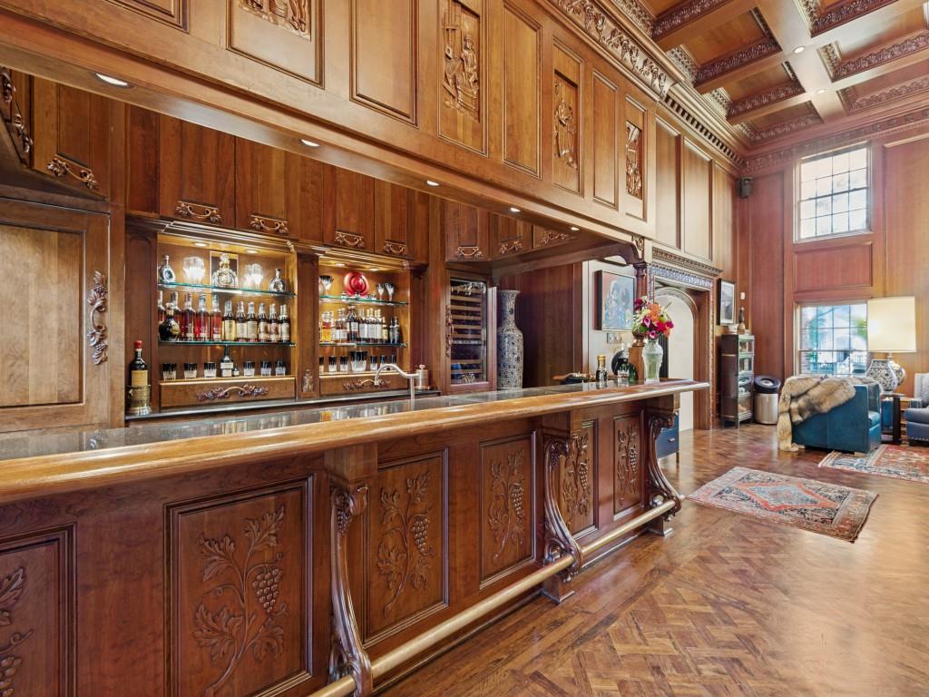 Custom wood-paneled bar featuring intricate relief carvings, coffered wood ceilings, built-in glass shelving, a metal foot rail, and herringbone-pattern wood flooring