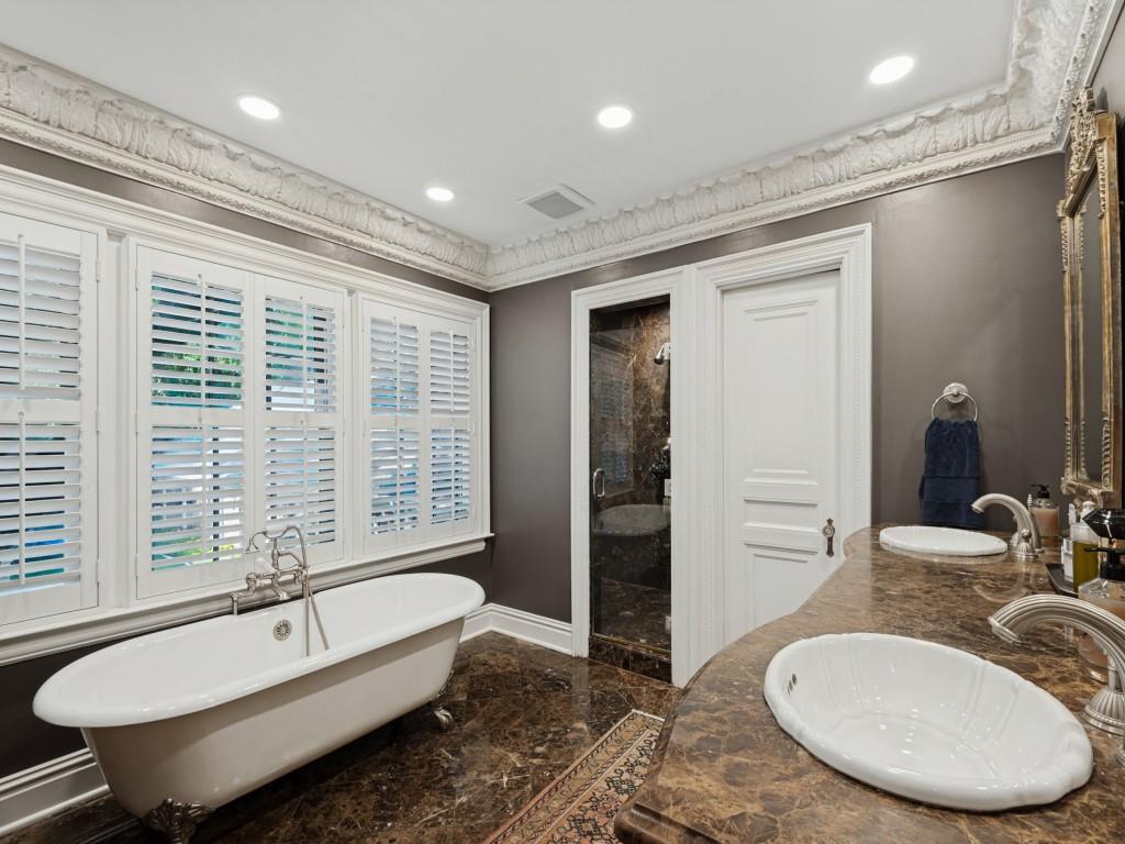 Freestanding clawfoot tub with bridge-style faucet, dual-sink vanity with stone countertop, walk-in stone-tiled shower, crown molding, and plantation shutters