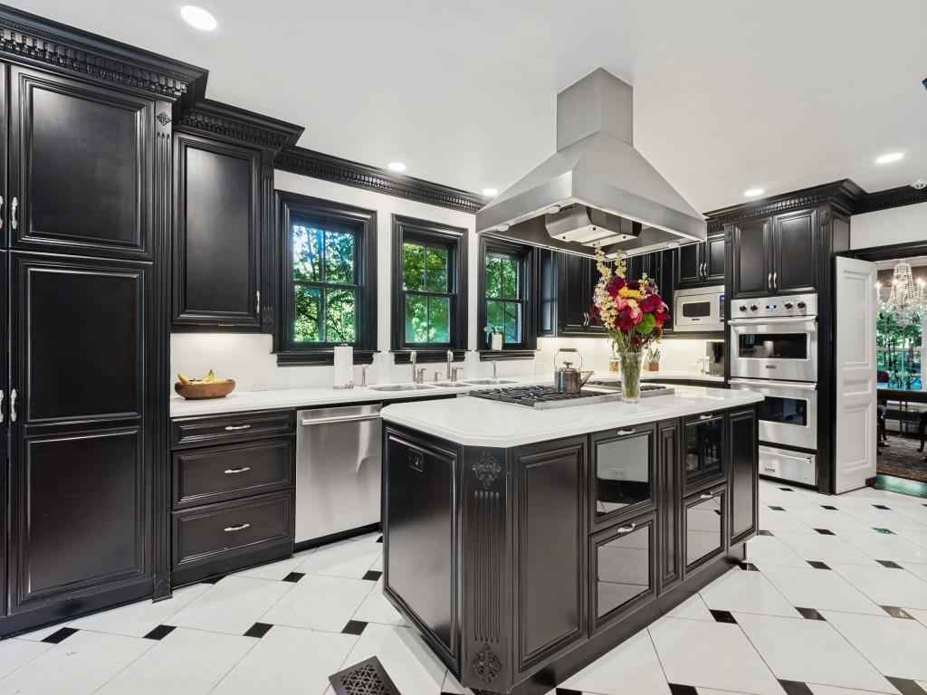 Custom dark cabinetry with ornate crown molding, expansive stone countertops, stainless steel appliances, a central island with a built-in gas cooktop, and polished tile flooring with decorative inlay