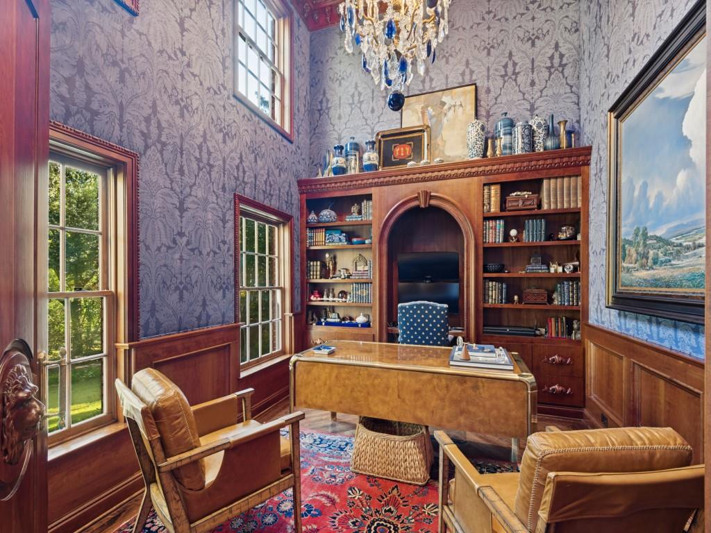 Study featuring custom floor-to-ceiling wood cabinetry with an arched niche, damask patterned wall coverings, wood wainscoting, multi-pane windows, and a tiered crystal chandelier