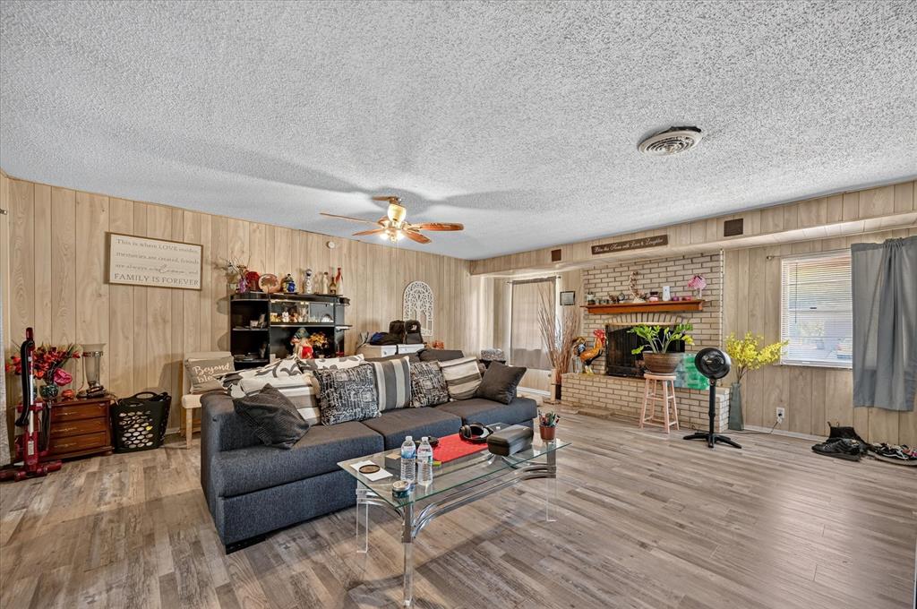 Expansive living area featuring wood-finish flooring, paneled wall accents, a brick-surround fireplace with a mantel, and an overhead ceiling fan