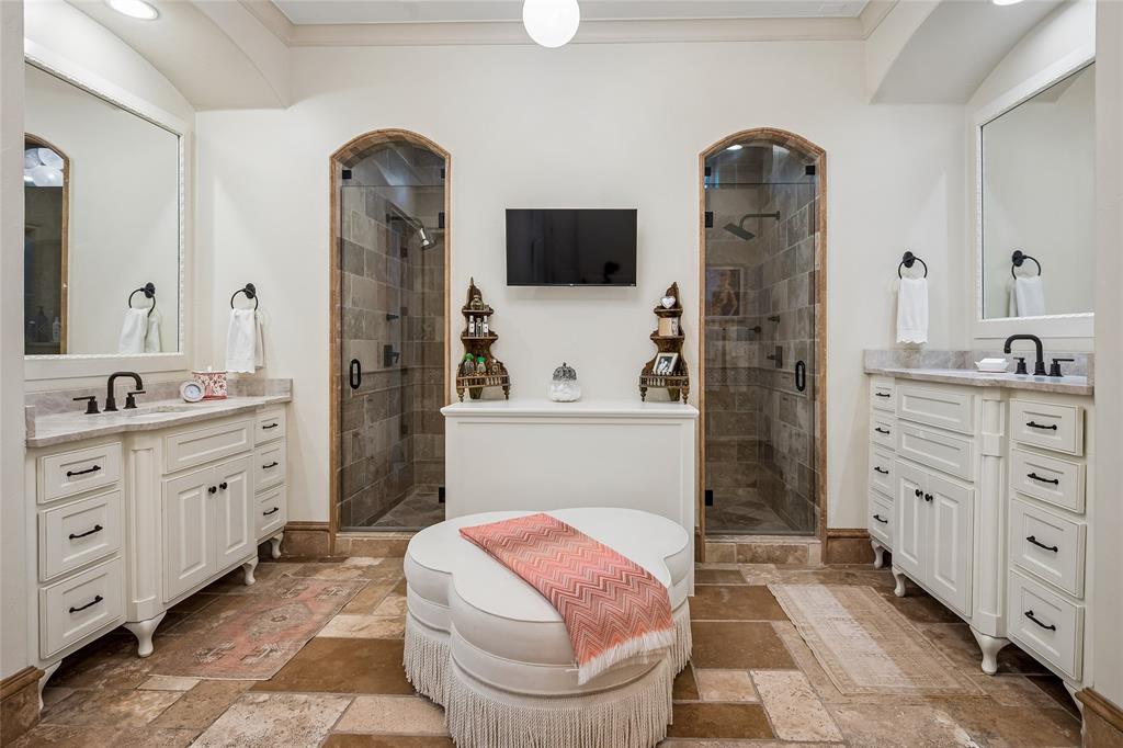 Primary bath offers dual vanity sinks with white cabinetry and framed mirrors, two glass-enclosed showers with tiled interiors, and stone-finish flooring