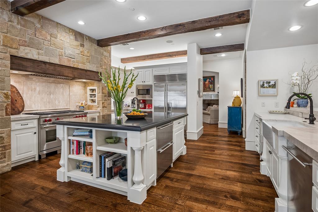 Gourmet kitchen featuring a stone accent wall, wood beams, stainless steel appliances, a center island with built-in shelving, and wood-finish flooring Gourmet kitchen featuring a stone accent wall, wood beams, stainless steel appliances, a center island with built-in shelving, and wood-finish flooring