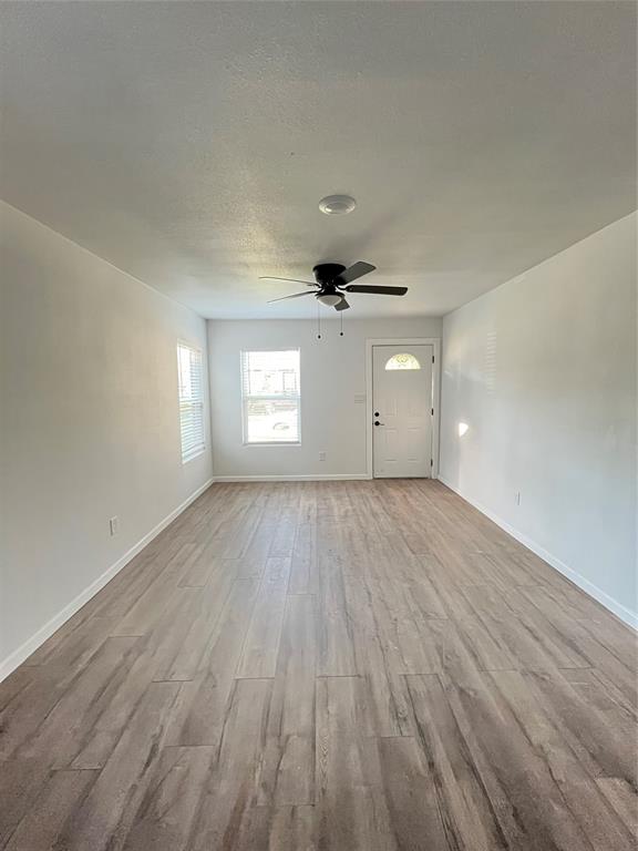 Neutral-toned room featuring wood-finish flooring, a ceiling fan with integrated lighting, dual windows, and an exterior door with a glass insert