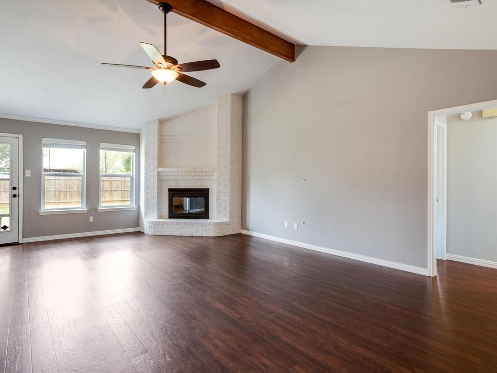 Vaulted ceiling featuring an exposed wood beam, corner white-painted brick fireplace, wood-finish flooring, and a ceiling fan with integrated light fixture