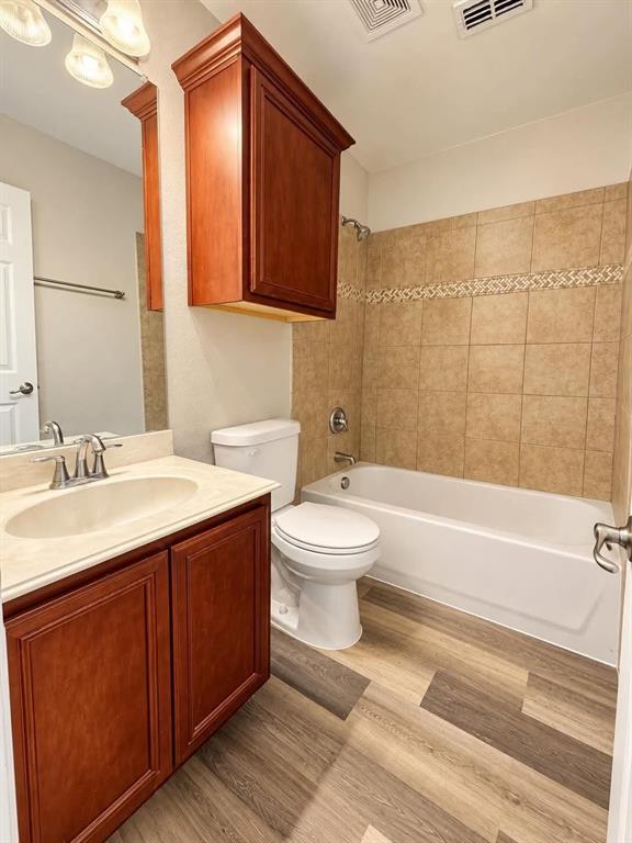 Wood-finish flooring, a vanity with a light-colored countertop and integrated sink, a built-in wood-toned storage cabinet, a bathtub with a tiled surround, and a toilet