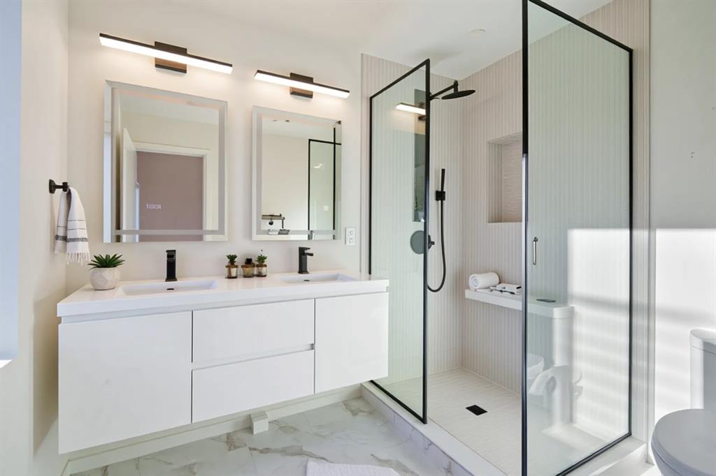 Dual floating vanity with integrated sinks and matte black fixtures, framed mirrors with overhead lighting, and a glass-enclosed walk-in shower with a built-in bench