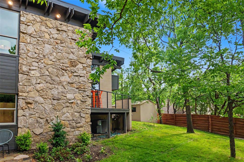 Multi-story exterior featuring natural stone masonry, elevated deck with horizontal rail system, large-pane windows, and landscaped grounds with mature trees and American cedar fencing