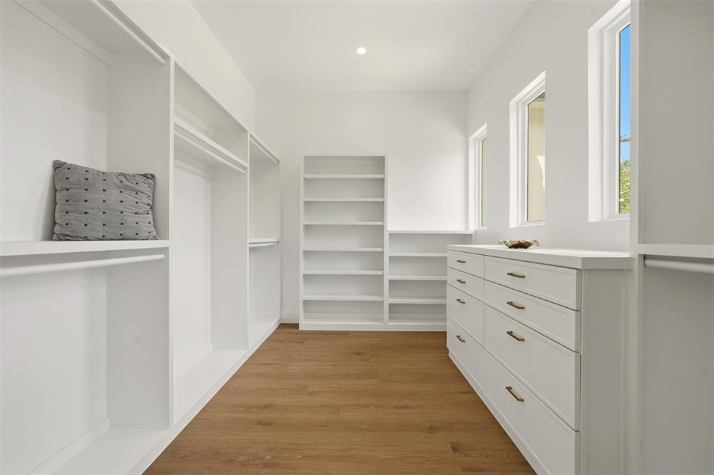 Spacious closet with wood-finish flooring, white built-in shelving, white built-in cabinetry with bronze hardware, and multiple windows