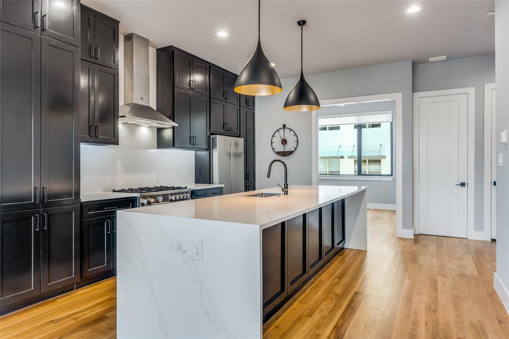 Open-concept kitchen featuring dark cabinetry, a gas range with stainless steel hood, a large island with a waterfall countertop, and wood-finish flooring