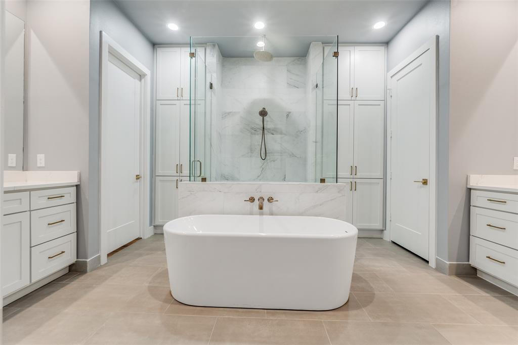 Freestanding soaking tub, dual vanities with stone countertops, glass-enclosed shower, built-in cabinetry, and tile flooring
