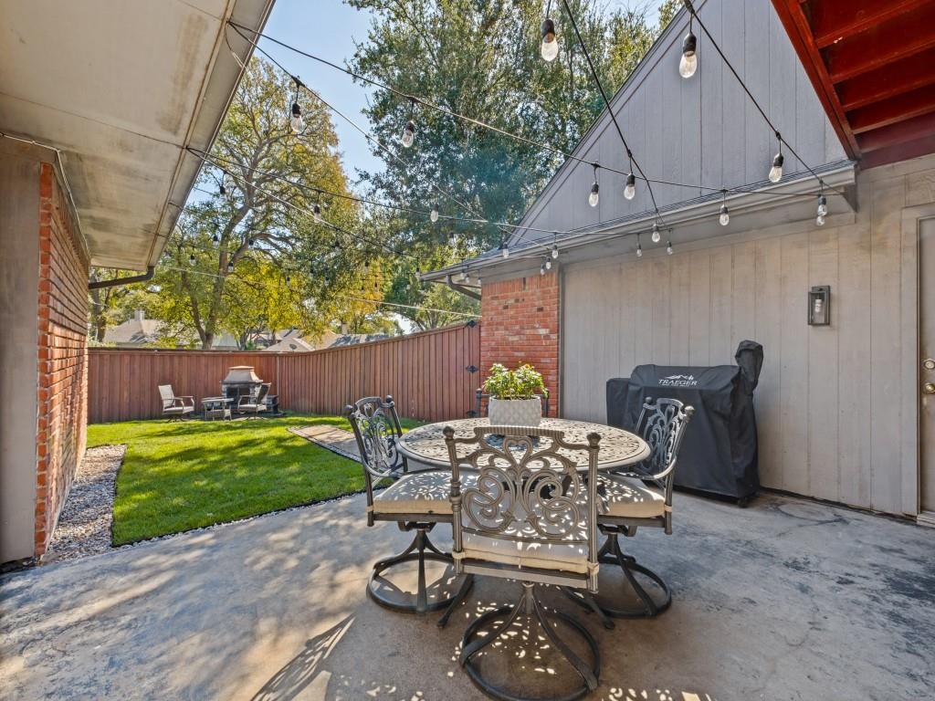Backyard patio featuring red brick accents, vertical siding, an attached wall-mounted lantern, and a wood privacy fence enclosing a lawn area with mature trees