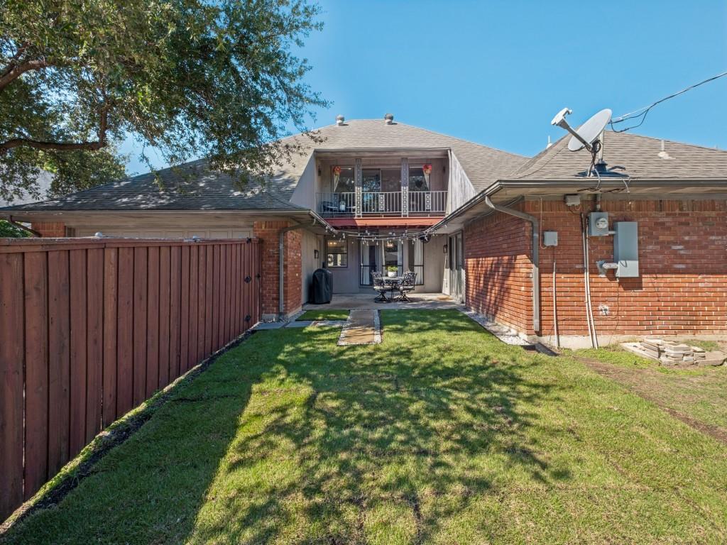 Brick exterior with a stone-paved patio, wooden privacy fencing, upper-level balcony with metal railing, and a grass lawn