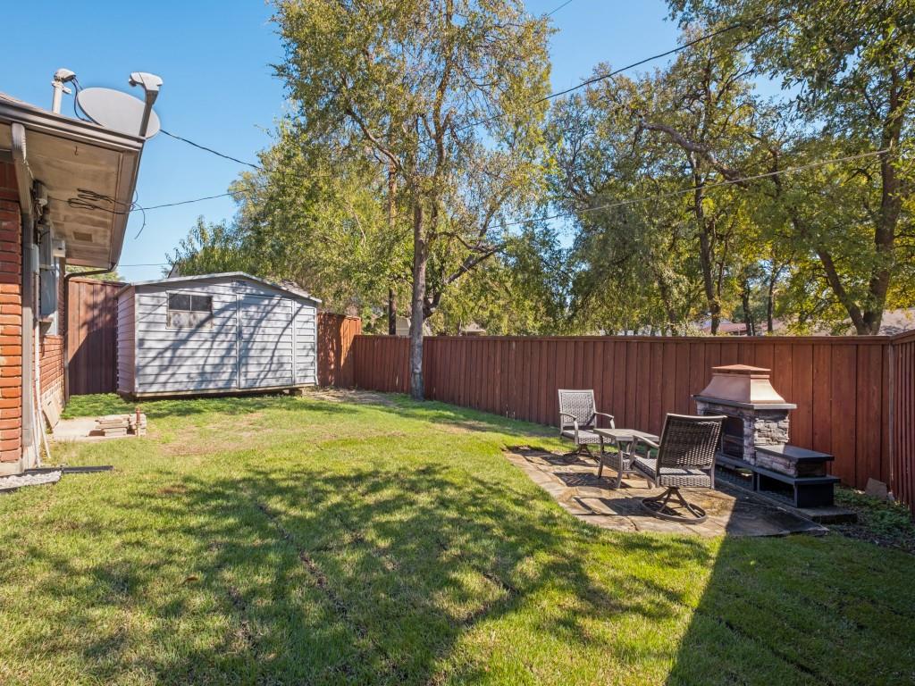 Backyard featuring a grass lawn, stone patio, stone-accented outdoor fireplace, storage shed, and wood-panel fencing