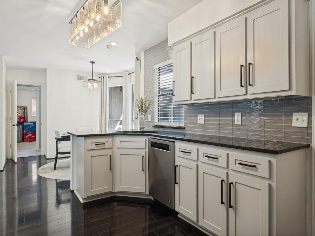 Kitchen featuring white cabinetry with matte black hardware, dark stone countertops, horizontal glass tile backsplash, wood-finish flooring, and a rectangular glass-paneled pendant light fixture Kitchen featuring white cabinetry with matte black hardware, dark stone countertops, horizontal glass tile backsplash, wood-finish flooring, and a rectangular glass-paneled pendant light fixture
