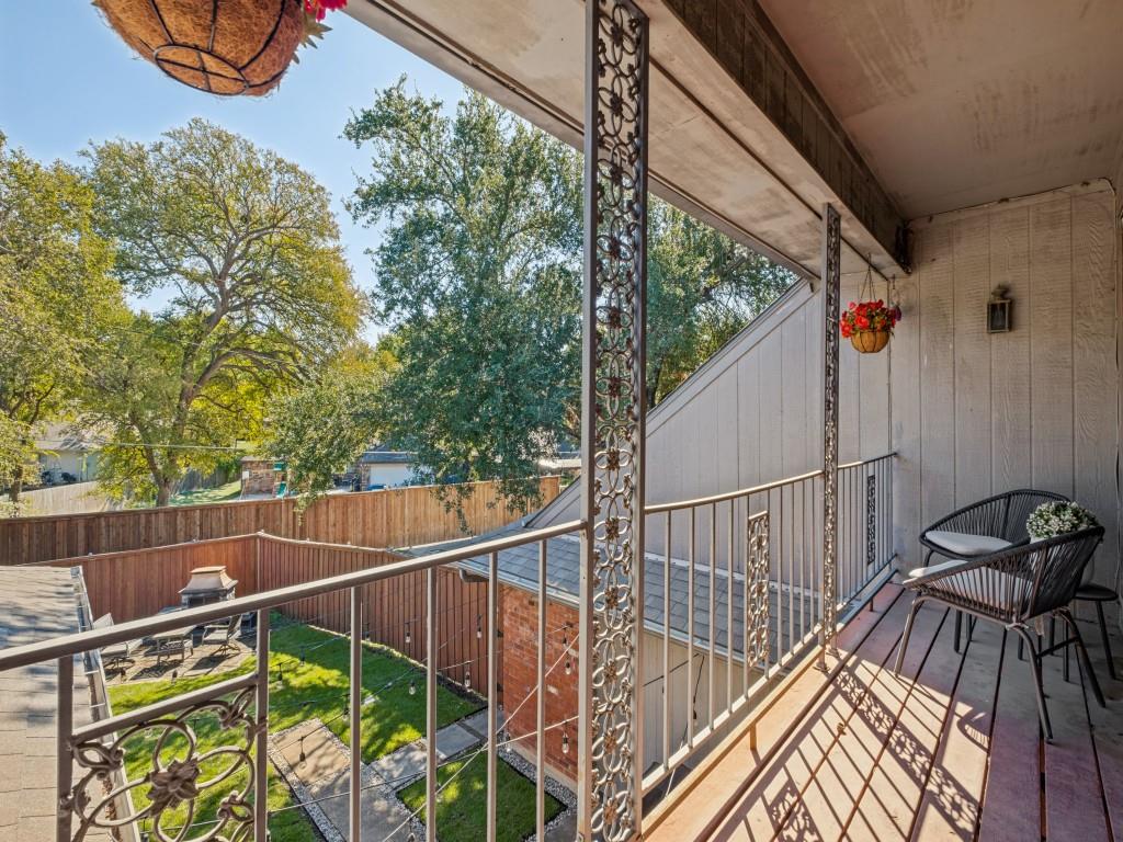 Covered balcony with decorative metal railings and vertical support columns overlooking a fenced backyard with mature trees