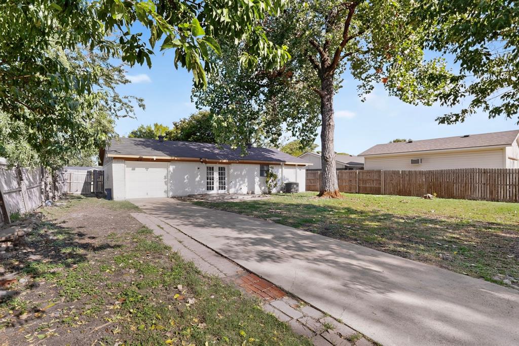 Exterior rear facade featuring a concrete driveway, multi-pane glass patio doors, mature trees, and a wood privacy fence