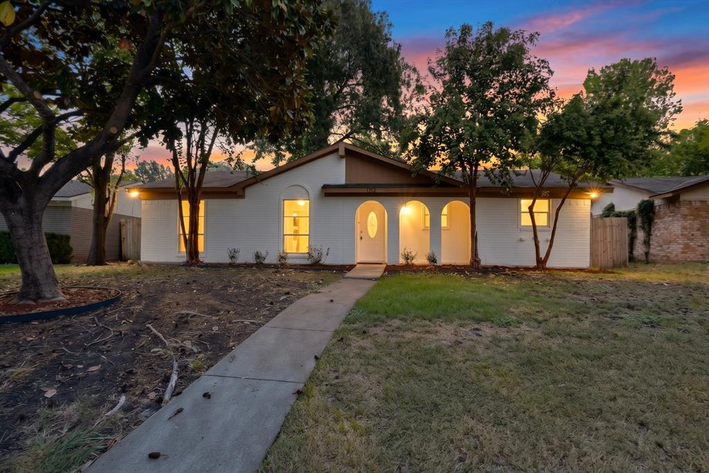 Single-story residence featuring white painted brick exterior, arched entryway, concrete walkway, and mature trees
