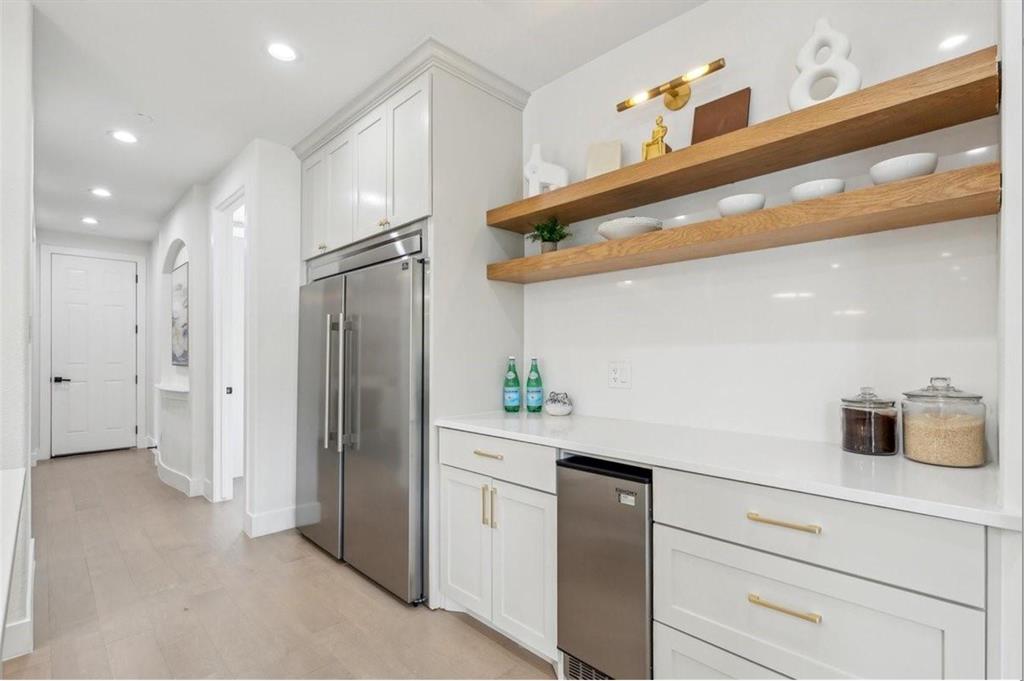 White cabinetry with gold-tone hardware, stainless steel refrigerator, built-in beverage cooler, white countertop, and wood-finish open shelving