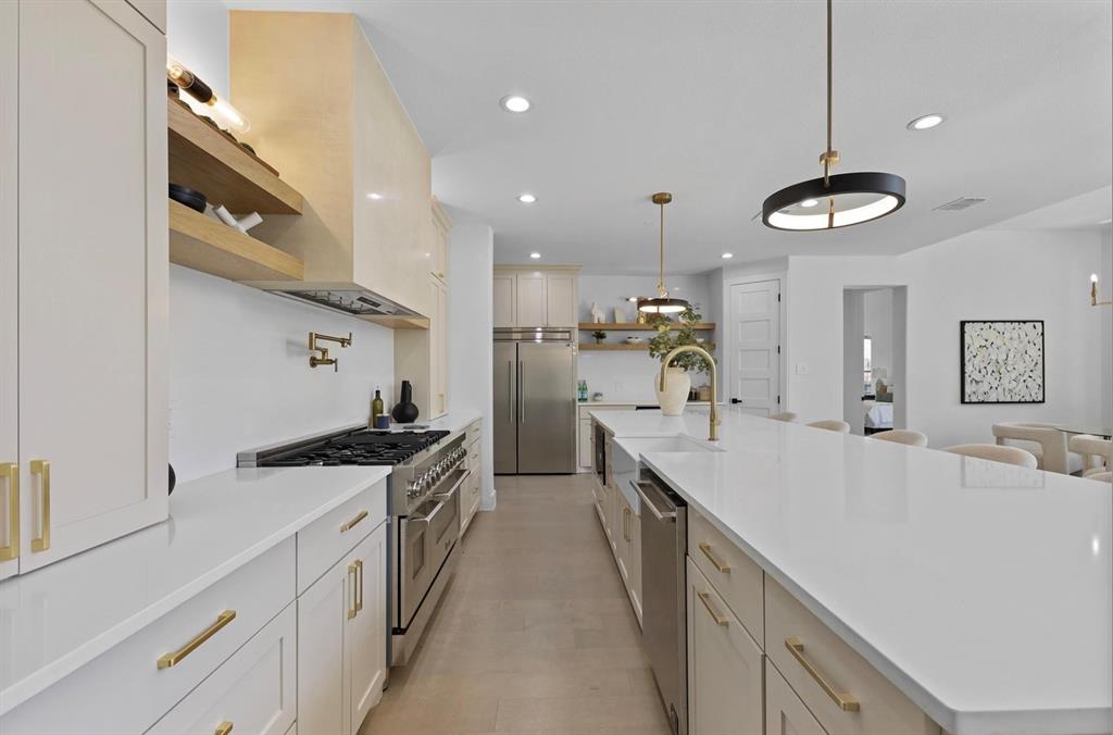 Expansive kitchen featuring a large central island with a sink, stainless steel appliances, white cabinetry with gold-finish hardware, and recessed lighting