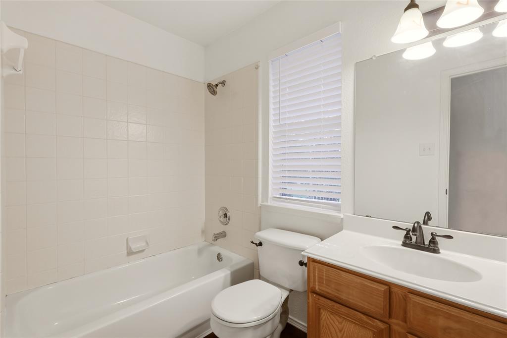 White tile bathtub surround, wood-finish vanity with white countertop, and window with privacy blinds