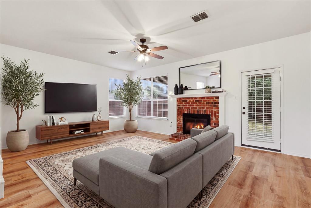 Open-concept living space with wood-finish flooring, a brick-surround fireplace with a white mantel, and ceiling fan.  Virtually staged.