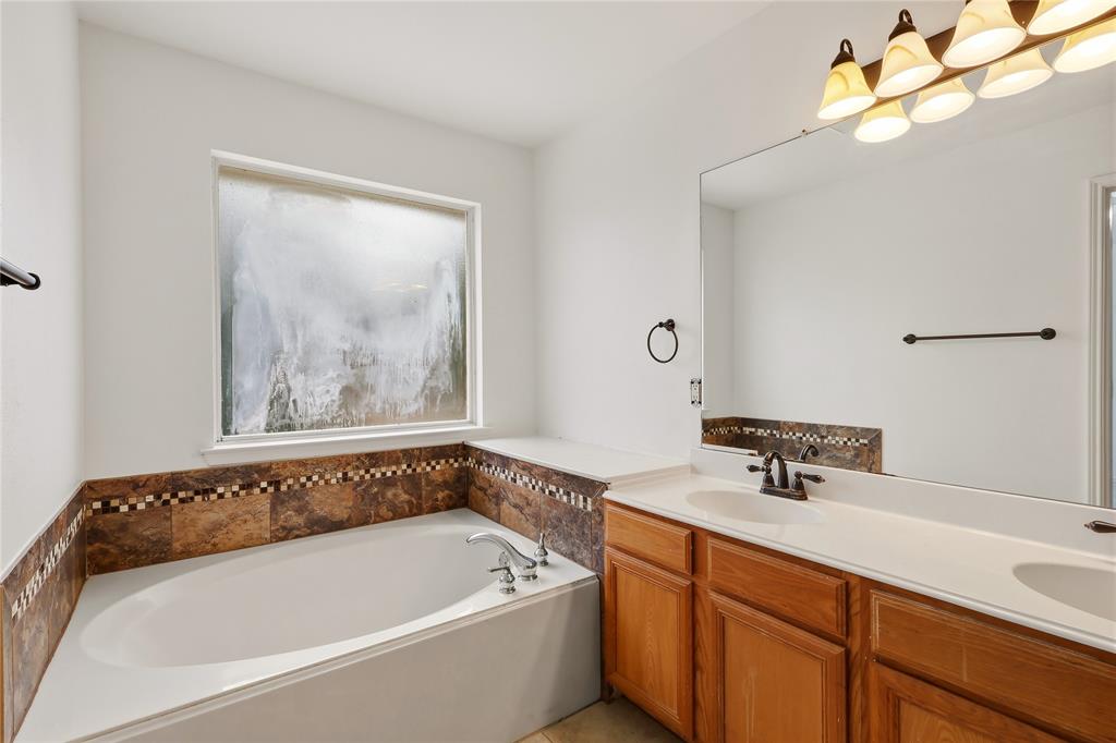 Built-in soaking tub, dual vanity with wood-finish cabinetry, extensive vanity mirror, wall-mounted light fixture with five sconces, and a tiled backsplash and tub surround