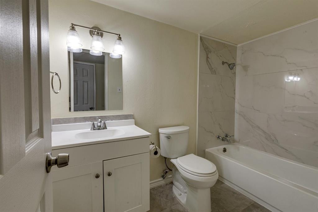 Bathroom featuring a white vanity cabinet with a solid-surface countertop, an integrated sink, three-light vanity fixture, and a shower-tub combination with full-height stone-finish wall tile