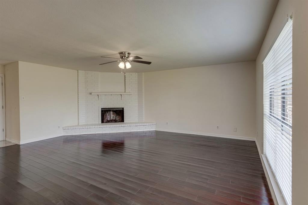 Spacious interior featuring wood-finish flooring, a corner fireplace with a brick surround, a mantel, and a ceiling fan with integrated lighting Spacious interior featuring wood-finish flooring, a corner fireplace with a brick surround, a mantel, and a ceiling fan with integrated lighting
