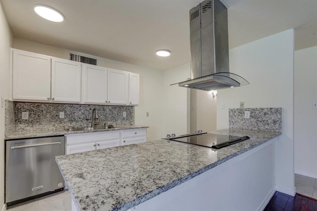 Kitchen featuring speckled stone countertops, white shaker-style cabinetry, stainless steel dishwasher, glass-accented stainless steel range hood, and a flat-surface cooktop Kitchen featuring speckled stone countertops, white shaker-style cabinetry, stainless steel dishwasher, glass-accented stainless steel range hood, and a flat-surface cooktop