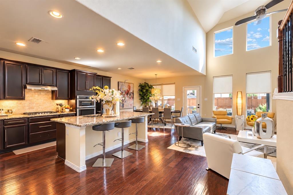 Open-concept kitchen and living area featuring dark cabinetry with stainless steel appliances, granite countertops, a large center island with seating, hardwood flooring throughout, and floor-to-ceiling windows with abundant natural light and skylight accents