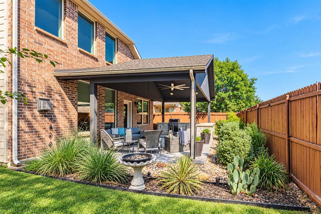 Covered patio with ceiling fan and integrated lighting overlooking a manicured lawn with ornamental grasses, desert plantings, and decorative stone hardscaping