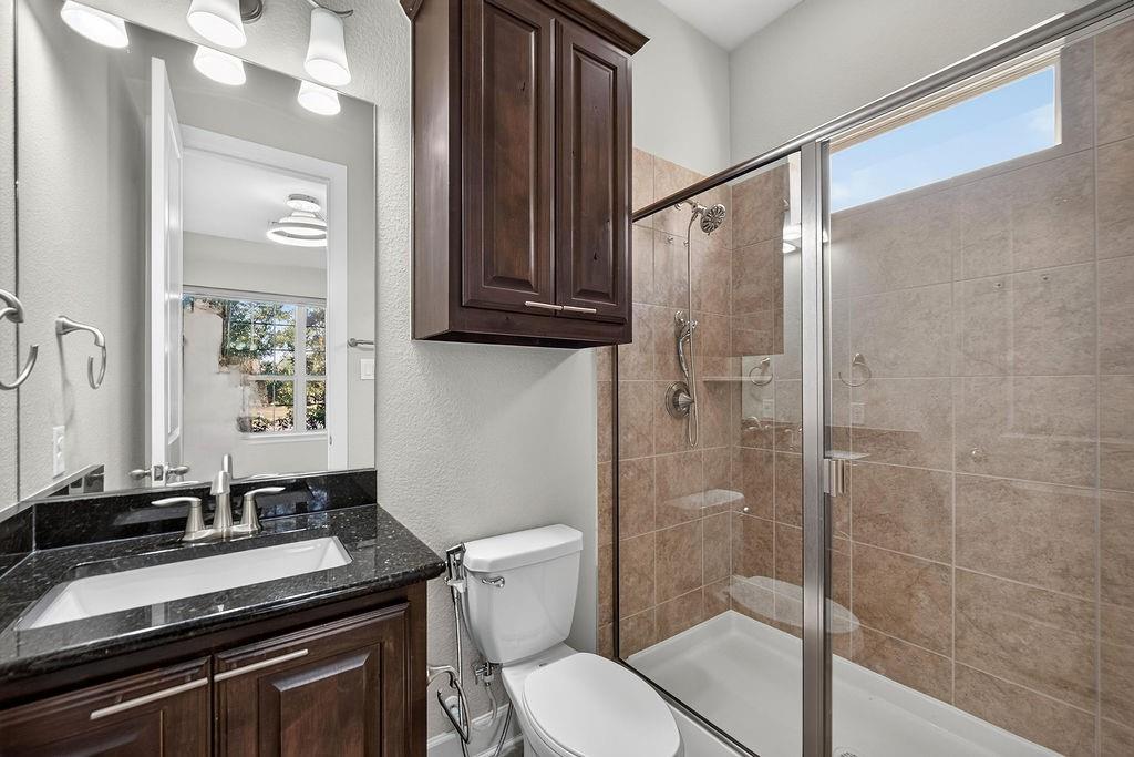 Dark-toned stone vanity countertop with an integrated rectangular sink, wall-mounted cabinetry, shower featuring tile surrounds and a transom window, and a glass-enclosed shower stall
