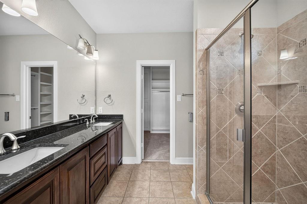 Dual-sink vanity with dark stone countertop, wood cabinetry, full-width mirror, and walk-in shower with diamond-pattern tile surround