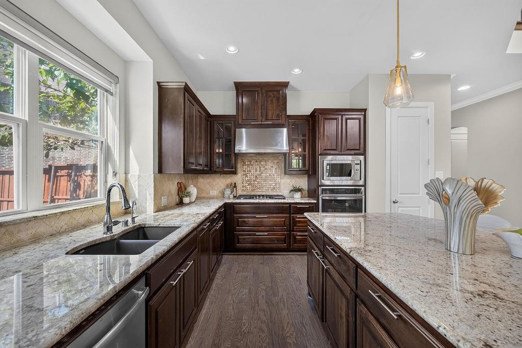Kitchen featuring dark wood cabinetry, expansive stone countertops, stainless steel appliances including a built-in oven and microwave, stone tile backsplash, and wood-finish flooring