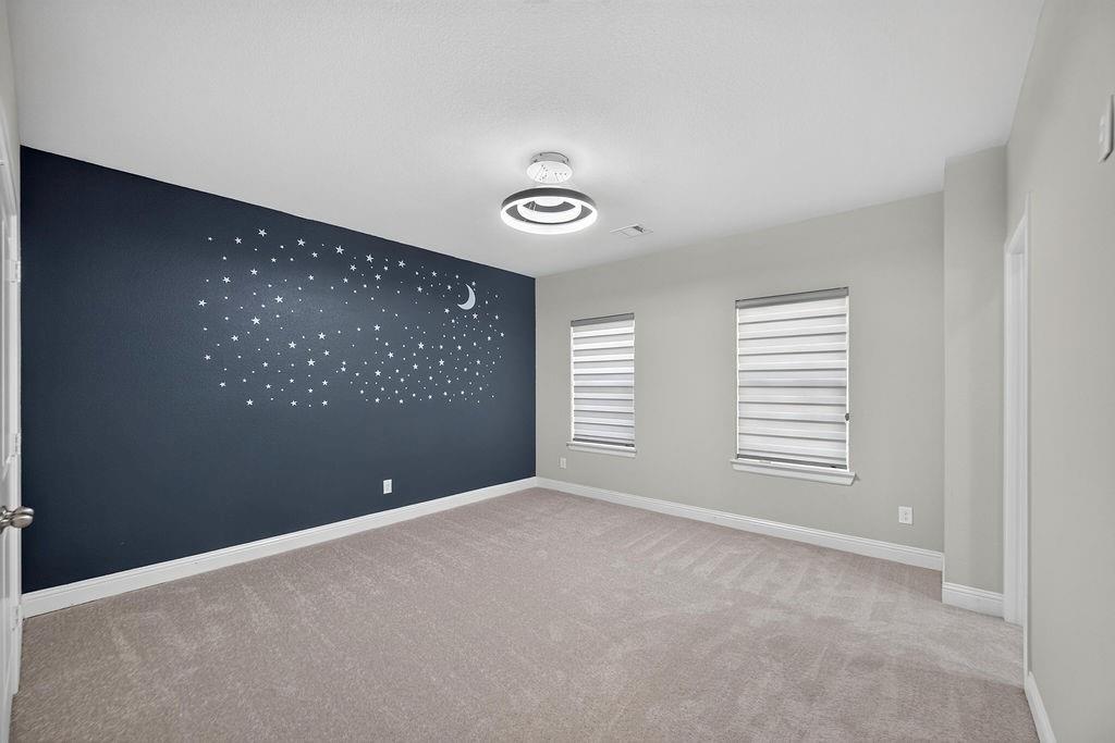 Neutral-toned room featuring wall-to-wall carpeting, a dark accent wall with mural detailing, dual windows with fitted light-filtering shades, and a modern circular LED light fixture