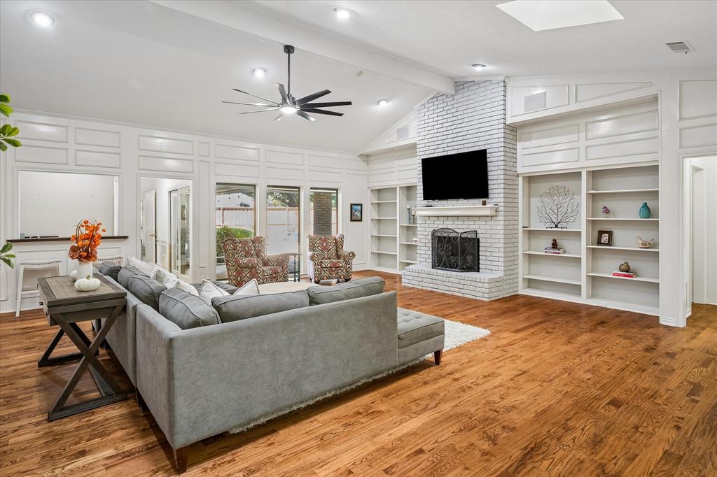 Vaulted ceiling with a contemporary ceiling fan, wood-finish flooring, white brick fireplace with a mantel, built-in shelving, and recessed lighting