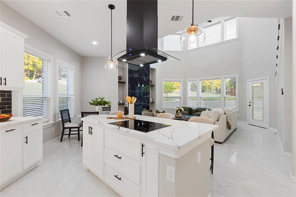 Open-concept living space with a kitchen island featuring an integrated cooktop, white cabinetry with dark hardware, a modern range hood, and high ceilings with upper windows