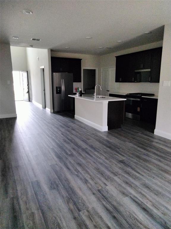 Open-concept kitchen featuring dark wood cabinetry, stainless steel appliances, a center island with sink, and light-toned countertops
