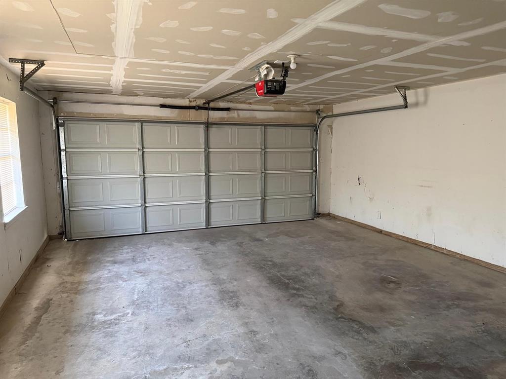 Concrete floor, white paneled garage door, exposed ceiling joists, window with blinds, garage door opener mechanism
