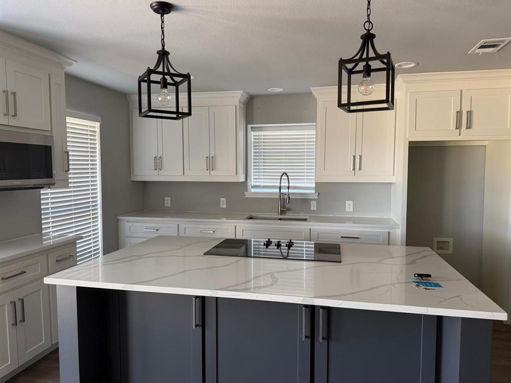 Kitchen featuring white shaker cabinetry, an island with a built-in cooktop, light-colored countertops with veining, a stainless steel microwave, and pendant lighting