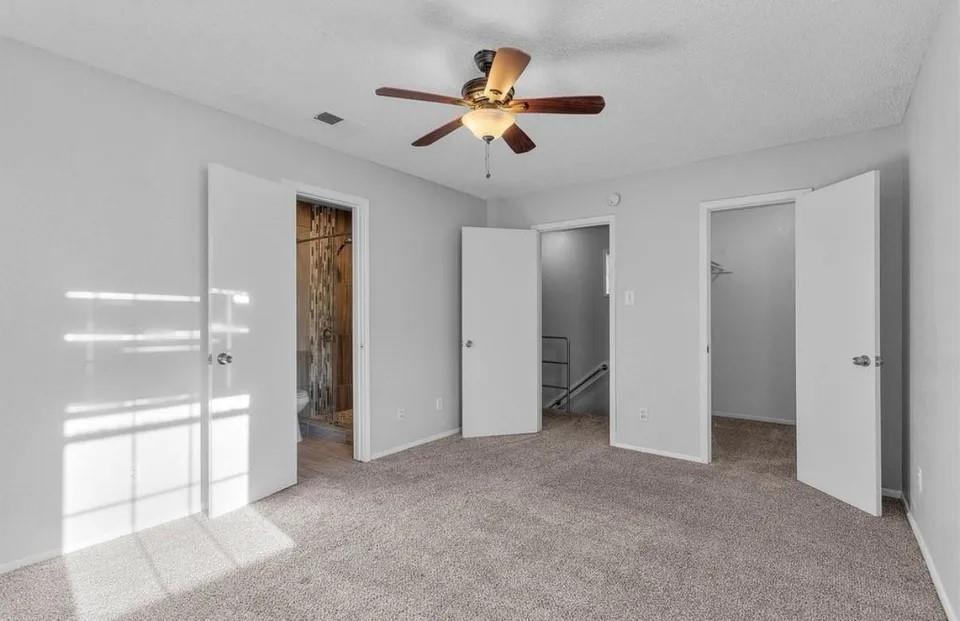 A spacious primary bedroom showcasing an en suite bathroom, built-in cabinet, ceiling fan and neutral-colored carpeting, perfect for relaxation and comfort.
